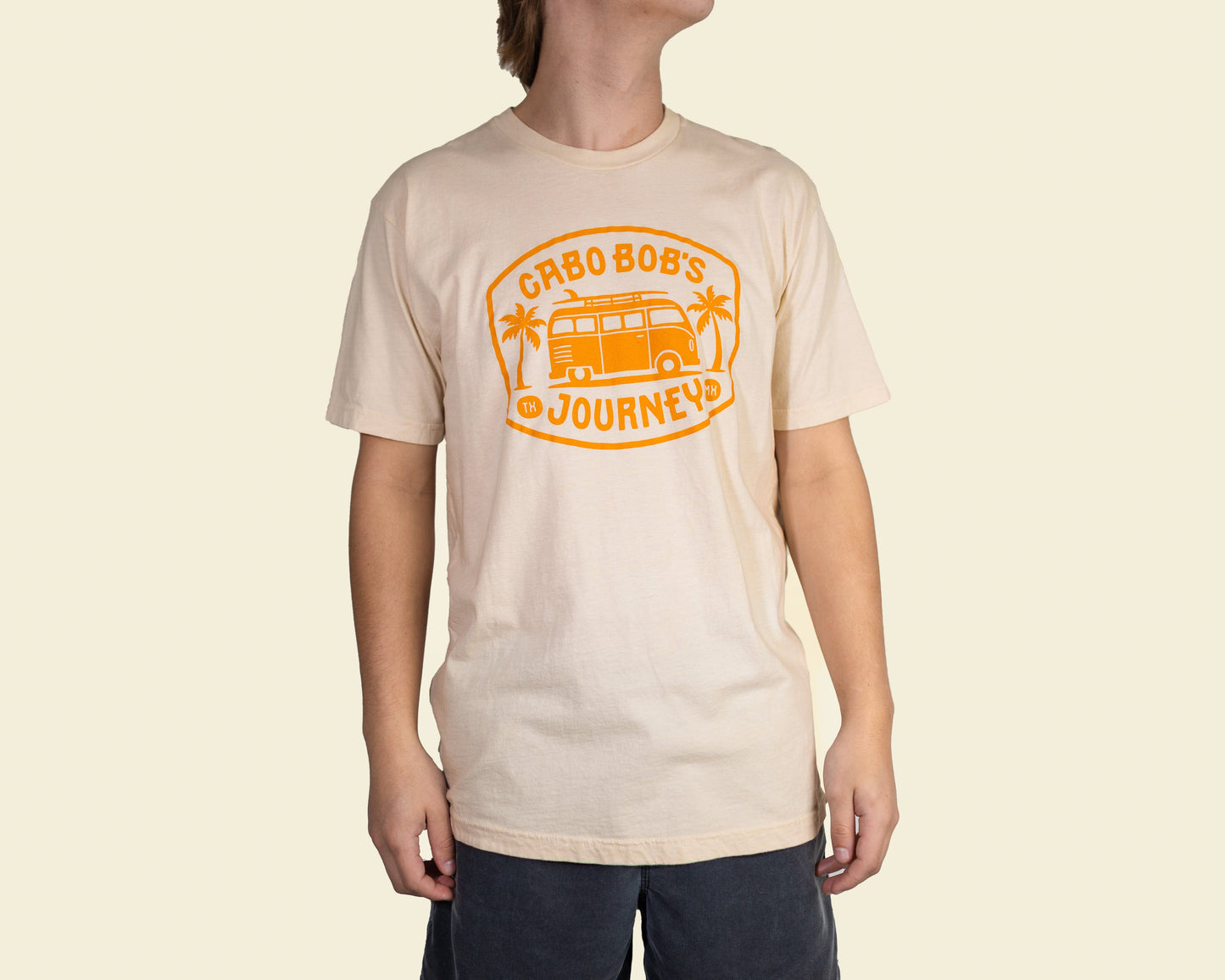 A person wearing a beige t-shirt with 'CABO BOB'S JOURNEYS' printed in orange on the front.